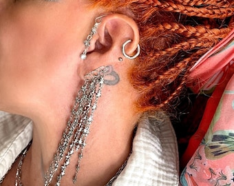 May include: A silver-toned ear cuff featuring a vertical chain design with small star-shaped charms that drape down from the earlobe. The jewelry piece includes a secondary small hoop earring and a stud, creating a layered metallic aesthetic.