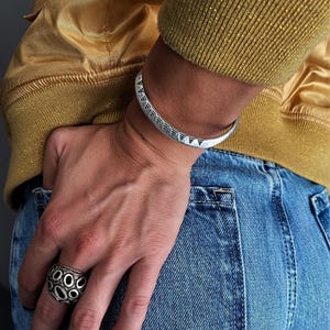 Pode incluir: Um bracelete de metal prateado com um padrão triangular repetitivo e detalhes escuros em baixo-relevo, combinado com um anel largo e texturizado de metal prateado com um design orgânico irregular de recortes circulares.