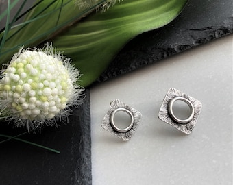 May include: A pair of silver-toned stud earrings featuring a square base with a textured, radiating line pattern and a smooth, raised circular center. The earrings have a metallic finish and a geometric design, showcasing a contrast between the textured square and the polished round element.