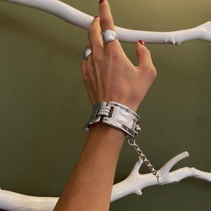 Könnte beinhalten: Ein breites, poliertes Armband in Silberoptik mit einem klappbaren Mittelglied-Design und einem hängenden Kettenanhänger, ergänzt durch zwei glatte, abgerundete Ringe in Silberoptik an den Fingern, alle mit einer reflektierenden Metalloberfläche.