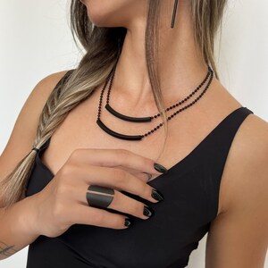 以下が含まれることがあります： マットブラックのジュエリーセット。カーブしたバーペンダント付きの二連ネックレス、長い直線的なドロップイヤリング、中央に縦の隙間がある幅広のオープンカフリングで構成されており、すべて滑らかで均一なダーク仕上げが施されています。