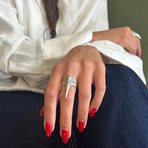 Pu&ograve; includere: Un anello color argento con un design ornato e un'estensione a punta. L'anello &egrave; indossato su un dito con smalto rosso. Lo sfondo mostra una camicia bianca e jeans blu.