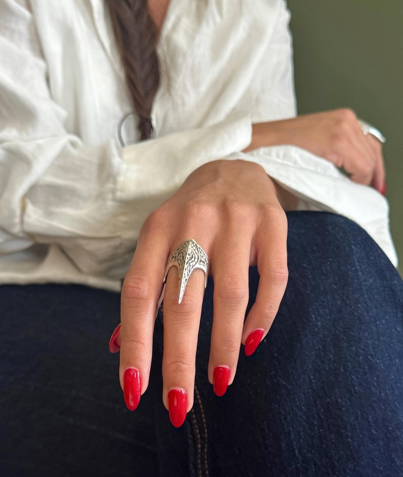 Pu&ograve; includere: Un anello color argento con un design ornato e un'estensione a punta. L'anello &egrave; indossato su un dito con smalto rosso. Lo sfondo mostra una camicia bianca e jeans blu.