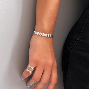 Puede incluir: Un brazalete plateado con una serie de cuentas circulares texturizadas. La imagen también muestra tres anillos plateados en los dedos. La persona lleva vaqueros negros y tiene esmalte de uñas azul claro.
