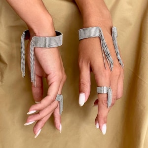 Könnte beinhalten: Ein Set aus zwei silberfarbenen Metallarmreifen und zwei passenden Ringen, jeweils mit einer feinen Netzstruktur und zarten, herabhängenden Kettenfransen. Die Schmuckstücke haben eine offene Struktur und zeichnen sich durch ein einheitliches metallisches Finish sowie detaillierte Gliederverbindungen aus.