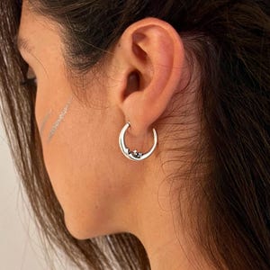 May include: A silver hoop earring shaped like a crescent moon with a detailed face embossed on the lower curve. The earring is polished and metallic, featuring a smooth finish that reflects light, worn in the earlobe with a simple circular design.