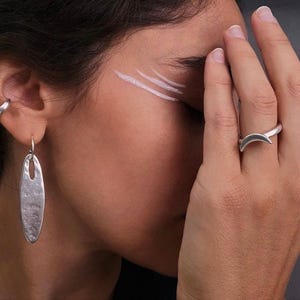 May include: A set of silver-toned jewelry consisting of a smooth ear cuff, a long oval-shaped drop earring with a hammered texture, a crescent moon ring, and a smooth oval-shaped ring. The pieces feature a matte finish and simple geometric shapes.