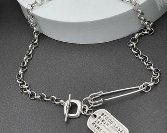 May include: A silver-toned rolo chain necklace featuring a toggle clasp, a large safety pin, and a rectangular dog tag pendant. The tag is engraved with the text NAME, TEL, BIRTH DATE, and BLOOD TYPE, each followed by a dotted line for personalization.