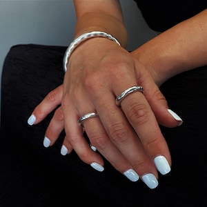 May include: A silver-toned metal bangle bracelet with a smooth, undulating surface, paired with two matching silver-toned rings worn on the fingers. The jewelry features a polished, reflective finish with a subtle, organic, and irregular shape.