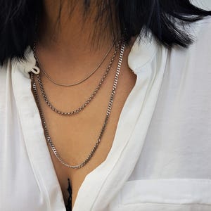 May include: A layered necklace set consisting of three distinct silver-toned chains. The top chain is a smooth, thin snake chain, the middle is a round cable link chain, and the bottom is a slightly thicker, flat curb chain, all worn together.