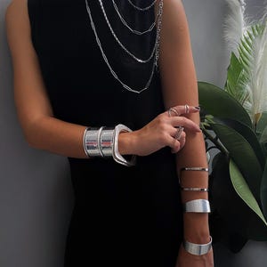 Op de afbeelding: Een mouwloze zwarte jurk gecombineerd met een zilverkleurige ketting met meerdere schakels die over de schouder valt. Het ensemble bevat verschillende brede zilverkleurige metalen armbanden om beide polsen en onderarmen, samen met meerdere zilverkleurige ringen.