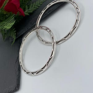 Op de afbeelding: Twee cirkelvormige zilverkleurige armbanden met een herhalend opengewerkt geometrisch patroon langs de buitenste band. De armbanden hebben een gepolijste metalen afwerking en een holle, lichtgewicht constructie met een constante breedte.