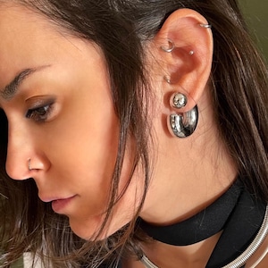 May include: A close-up of an ear with multiple piercings, including a large, shiny silver hoop earring. The earring has a rounded, open design. The person is wearing a black top and a silver necklace.