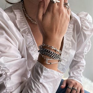 May include: A collection of silver-toned jewelry including a barbed wire chain necklace, a layered bracelet set featuring a safety pin charm and curb chains, and multiple rings including a cross design and a studded band, worn with a white blouse.