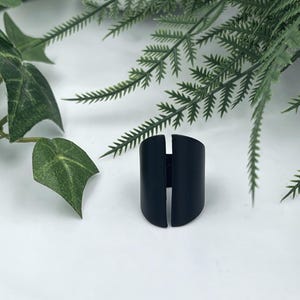 以下が含まれることがあります： 中央に垂直なスリットが入った幅広でカーブしたバンドを持つマットブラックのリング。指の裏側で合わさる2つの独立した対称的な半分で構成されています。表面は滑らかで光沢のない仕上げになっています。