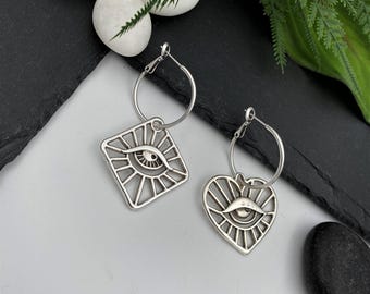 May include: A pair of silver-toned hoop earrings featuring dangling charms. One charm is a diamond shape and the other is a heart shape, both containing a cut-out eye design with radiating lines extending from the center to the outer edges.