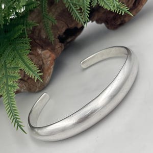 May include: A solid silver-toned cuff bracelet with a smooth, rounded surface and an open-ended design. The bracelet features a polished, reflective finish that catches the light, showcasing a simple, curved shape that tapers slightly at both ends.