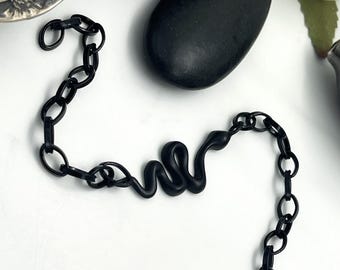 May include: A matte black chain bracelet featuring a central snake-shaped link. The bracelet is constructed from oval-shaped links and includes a lobster claw clasp at one end, providing a secure closure for this dark, monochromatic accessory.