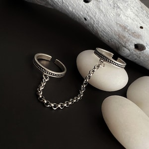 Op de afbeelding: Een zilverkleurige dubbele ring verbonden door een fijne metalen ketting. Elke ring heeft een open ontwerp met een textuur van kraaltjes langs de buitenrand. Het sieraad is gemaakt van gepolijst metaal met een licht geoxideerde afwerking.