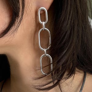 May include: A long drop earring featuring three interconnected silver-toned oval links. The earring consists of a small top link attached to a medium middle link, which connects to a larger bottom link, creating a vertical chain-like structure.