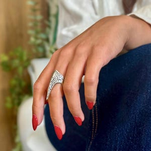 Pu&ograve; includere: Un anello color argento con un design intricato e inciso. L'anello ha una forma triangolare con un'estremit&agrave; a punta. La mano che indossa l'anello ha lo smalto rosso e poggia su una gamba vestita di denim blu.