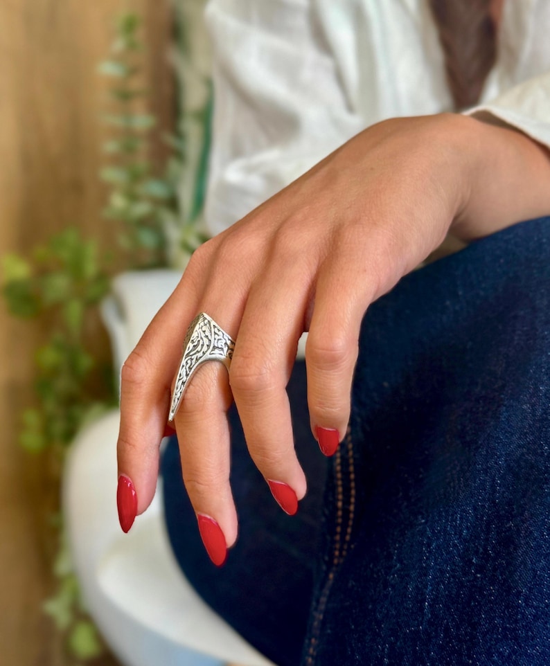 Pu&ograve; includere: Un anello color argento con un design intricato e inciso. L'anello ha una forma triangolare con un'estremit&agrave; a punta. La mano che indossa l'anello ha lo smalto rosso e poggia su una gamba vestita di denim blu.