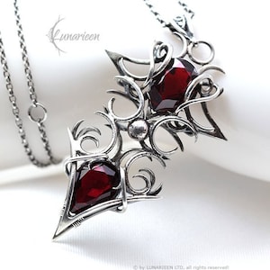 Könnte beinhalten: Eine silberne Halskette mit einem detaillierten, komplizierten Design mit zwei roten Edelsteinen. Der Anhänger ist in Form einer stilisierten Pfeilspitze mit einer spitzen Spitze gestaltet.