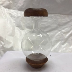 May include: A clear glass vase with a round body and a wooden lid. The vase has a wide opening at the top and a narrow neck.