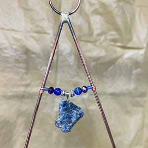 May include: A handmade pendant featuring a rough-cut blue stone suspended from a copper-colored metal frame. The pendant includes blue beads and wire accents. The frame has a triangular shape with a circular top, suitable for hanging.