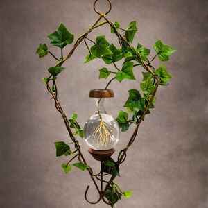 May include: A hanging planter featuring a diamond-shaped bronze metal frame entwined with green artificial ivy vines. A clear glass bulb-shaped vase is suspended in the center, containing a plant with visible yellow roots submerged in water.