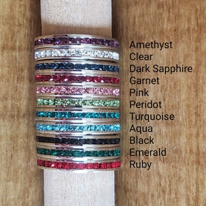 May include: Stack of 12 silver rings with different colored crystal stones. The stones are amethyst, clear, dark sapphire, garnet, pink, peridot, turquoise, aqua, black, emerald, and ruby.