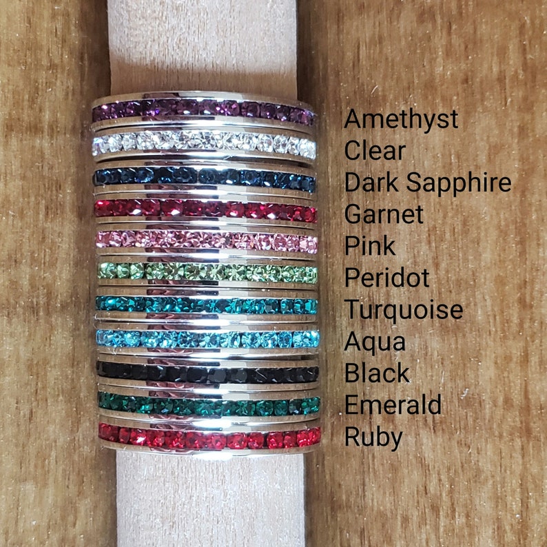 May include: Stack of 12 silver rings with different colored crystal stones. The stones are amethyst, clear, dark sapphire, garnet, pink, peridot, turquoise, aqua, black, emerald, and ruby.