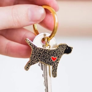 May include: A gold-colored keychain with a black and gold dog charm. The dog charm has a red heart and is attached to a key. The keychain is held by a hand.