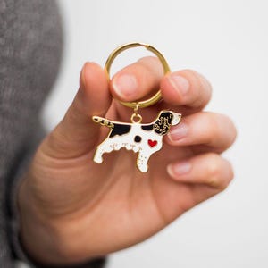 May include: A gold-colored metal dog keychain with a white body, black spots, and a red heart. The dog-shaped charm is attached to a gold ring. The keychain is held in a person's hand.