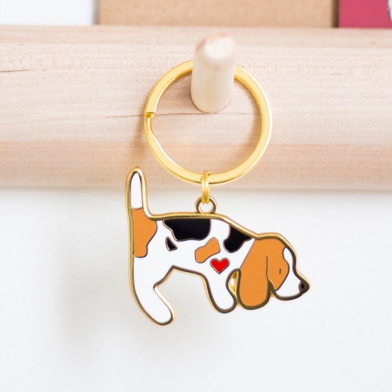 Beagle dog gold plated enamel keyring.  White, black and tan coloured enamel with a little black nose, a red heart on the chest, gold plated lines and sand blasted on the reverse side. Dog keyring is attached to a gold plated split keyring hoop.