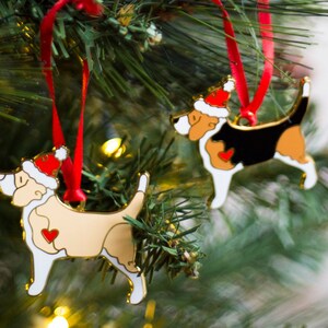 Puede incluir: Dos adornos de perro de color dorado con gorros de Pap&aacute; Noel rojos colgados de un &aacute;rbol de Navidad. Los adornos son blancos, negros y marrones con corazones rojos en el pecho.