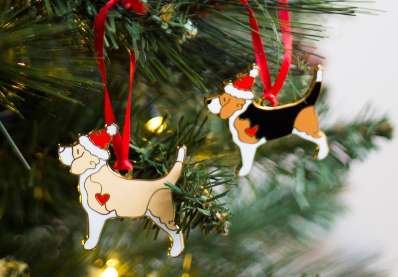 Puede incluir: Dos adornos de perro de color dorado con gorros de Pap&aacute; Noel rojos colgados de un &aacute;rbol de Navidad. Los adornos son blancos, negros y marrones con corazones rojos en el pecho.
