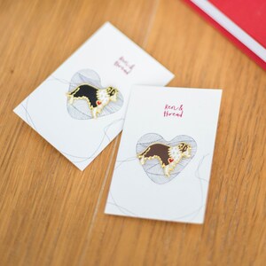 A pair of border collie gold plated enamel pin badges on backing cards. One Brown and white Border collie dog, one black and white Border collie sheep dog. Each pin features Ren's trademark little red heart on it's chest.