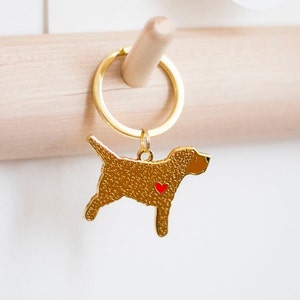 May include: Gold-colored dog-shaped keychain with a red heart detail. The keychain is attached to a gold ring and hangs from a wooden dowel. The dog charm has a textured surface.