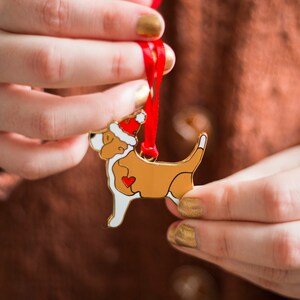 Puede incluir: Un adorno de metal dorado en forma de perro con un gorro de Pap&aacute; Noel rojo. El perro sostiene un coraz&oacute;n rojo en sus patas. El adorno est&aacute; colgado de una cinta roja.