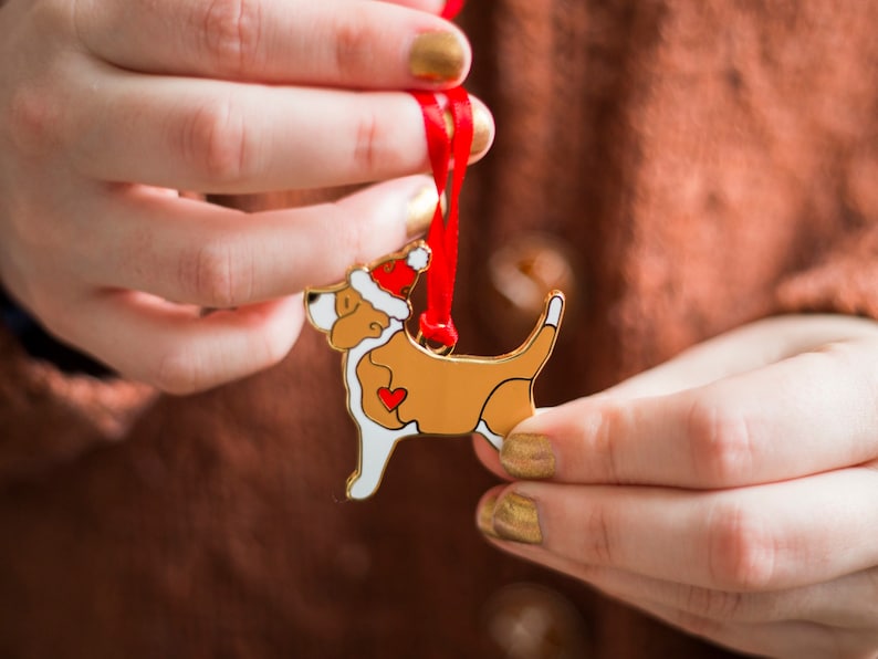 Puede incluir: Un adorno de metal dorado en forma de perro con un gorro de Pap&aacute; Noel rojo. El perro sostiene un coraz&oacute;n rojo en sus patas. El adorno est&aacute; colgado de una cinta roja.