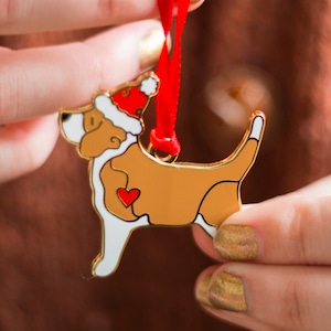 Puede incluir: Adorno navide&ntilde;o de metal dorado en forma de perro con un gorro de Pap&aacute; Noel rojo. El perro tiene un coraz&oacute;n blanco en el pecho.