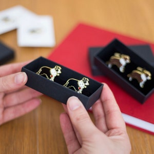May include: Two black and white border collie dog pins in a black box. The pins are gold-toned and have red hearts on their chests.