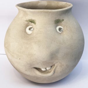 Könnte beinhalten: Eine skurrile, handgefertigte Keramikvase mit einem lächelnden Gesicht. Die Vase hat eine hellbeige Farbe mit bemalten weißen Augen, grünen Augenbrauen und einem lächelnden Mund. Sie hat einen runden Körper und eine weite Öffnung oben.