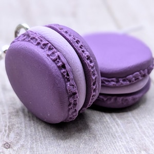 Könnte beinhalten: Nahaufnahme von zwei lila Makronen, eine davon mit einem silbernen Schlüsselanhänger. Die Makronen haben einen strukturierten, geriffelten Rand und eine hellere lilafarbene Füllung. Der Hintergrund ist eine hellgraue Holzoberfläche.