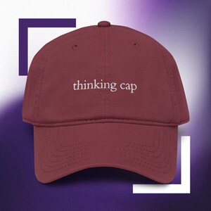 Op de afbeelding: Een kastanjebruine baseballpet met de tekst "thinking cap" geborduurd in wit garen op het voorpaneel. De pet heeft een gebogen klep, een knoopje aan de bovenkant en gestikte oogjes voor ventilatie op de panelen van de bol.