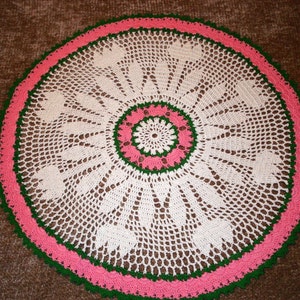 May include: White crocheted doily with a green and pink border. The doily has a circular design with a center that is a smaller circle with a pink and green border.