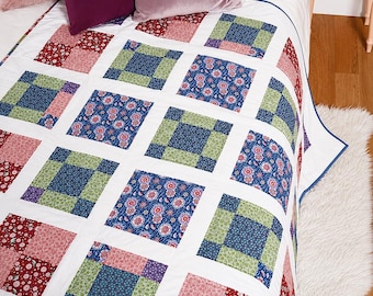 May include: A patchwork quilt featuring a repeating grid pattern of white sashing separating square blocks. The blocks alternate between floral prints in shades of red, blue, and green, and solid-coloured squares in deep blue and light green.