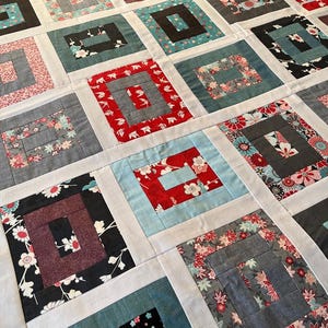 May include: A patchwork quilt featuring a repeating log cabin block pattern. Each square consists of various fabric prints in shades of red, black, blue, grey, and white, separated by solid white sashing strips that form a grid across the surface.