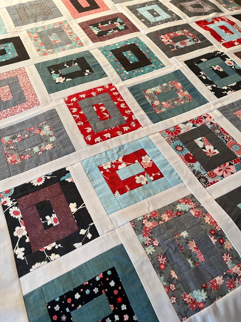 May include: A patchwork quilt featuring a repeating log cabin block pattern. Each square consists of various fabric prints in shades of red, black, blue, grey, and white, separated by solid white sashing strips that form a grid across the surface.